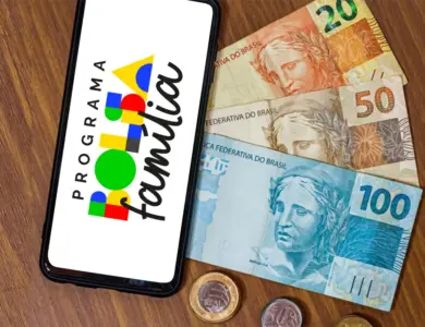 Smartphone exibindo logo Bolsa Família ao lado de notas e moedas de real sobre mesa de madeira.