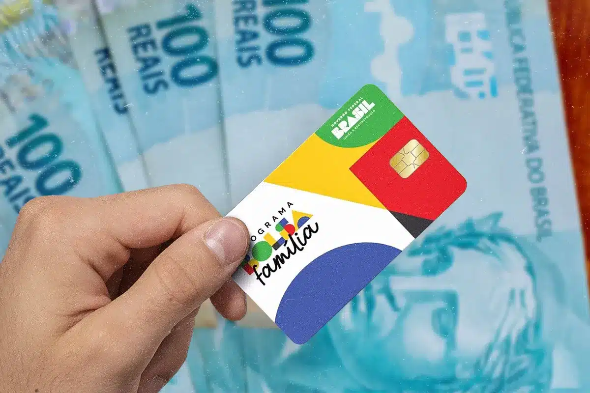 Cartão do Bolsa Família segurado por uma pessoa, com notas de real ao fundo, representando o pagamento antecipado do benefício em cidades de Minas Gerais.