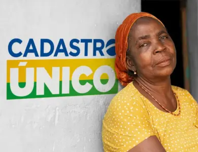 Mulher de lenço laranja e blusa amarela encostada em parede com logo do Cadastro Único ao fundo