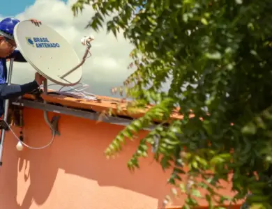 Técnico com capacete azul instala antena parabólica digital do programa Brasil Antenado no telhado de casa cor-de-rosa.