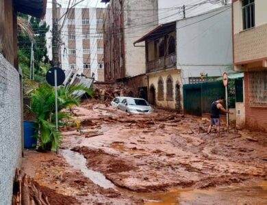 Rua coberta por lama após enchente com carro parcialmente submerso e homem caminhando em meio à destruição