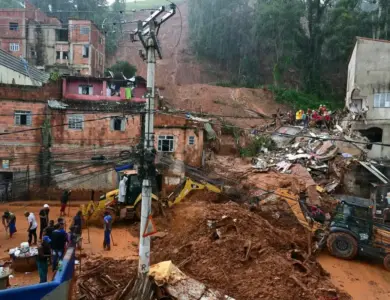 Retroescavadeiras amarelas removem lama de deslizamento em comunidade com casas de tijolos vermelhos enquanto moradores observam.