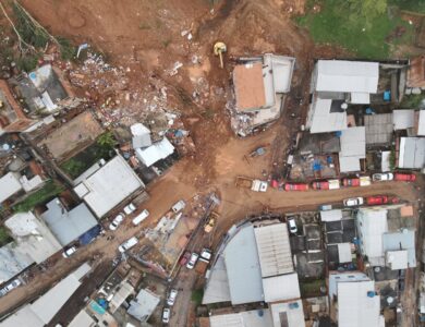 Vista aérea de deslizamento de terra atingindo casas em bairro residencial com escavadeiras e equipes de resgate no local.