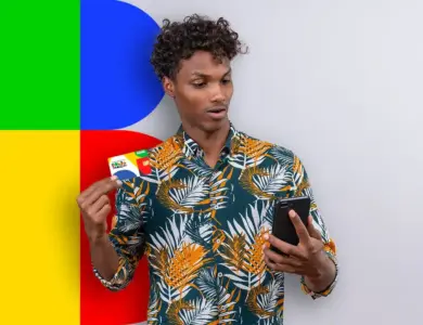 Jovem negro de cabelos cacheados, camisa tropical verde e laranja, segura cartão Bolsa Família e olha celular preto.