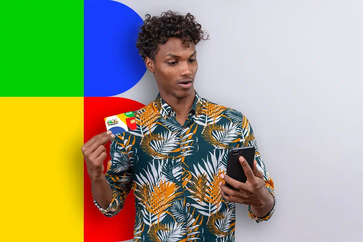 Jovem negro de cabelos cacheados, camisa tropical verde e laranja, segura cartão Bolsa Família e olha celular preto.