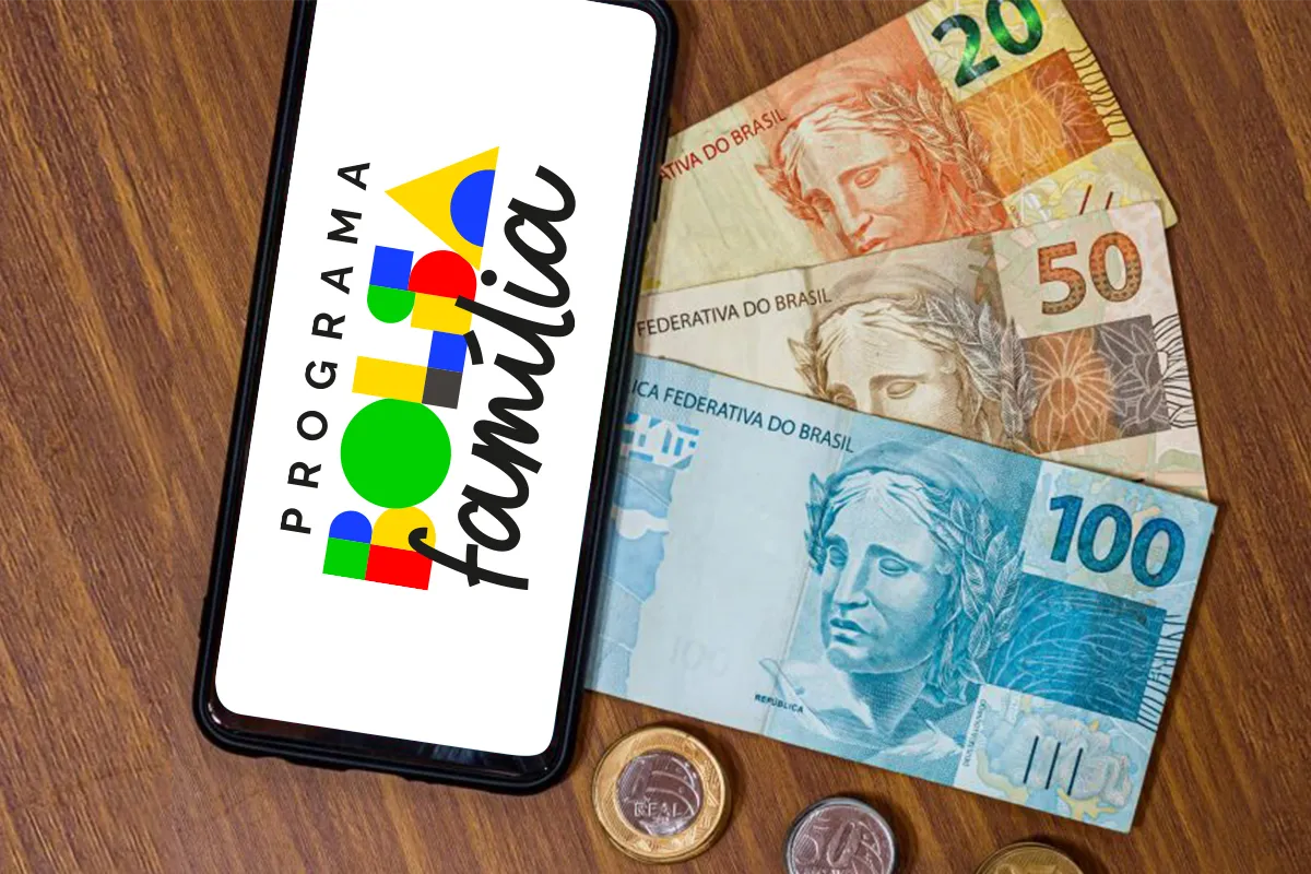 Celular com a logomarca do Programa Bolsa Família exibida na tela, ao lado de notas e moedas de reais sobre uma mesa, representando a consulta ao pagamento antecipado do benefício pelo aplicativo em abril de 2026