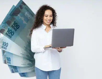 Mulher jovem sorrindo segura notebook cinza com notas de 100 reais ao fundo em fundo branco.