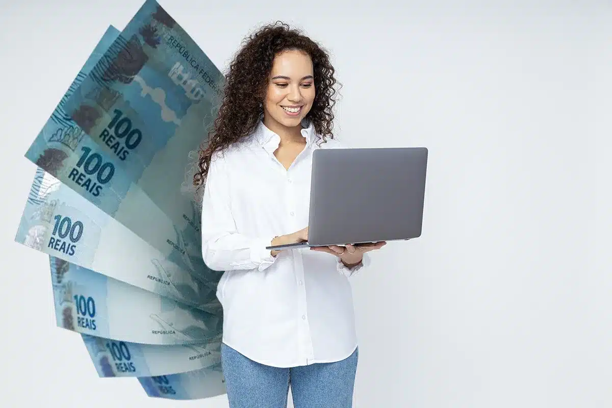 Mulher jovem sorrindo segura notebook cinza com notas de 100 reais ao fundo em fundo branco.