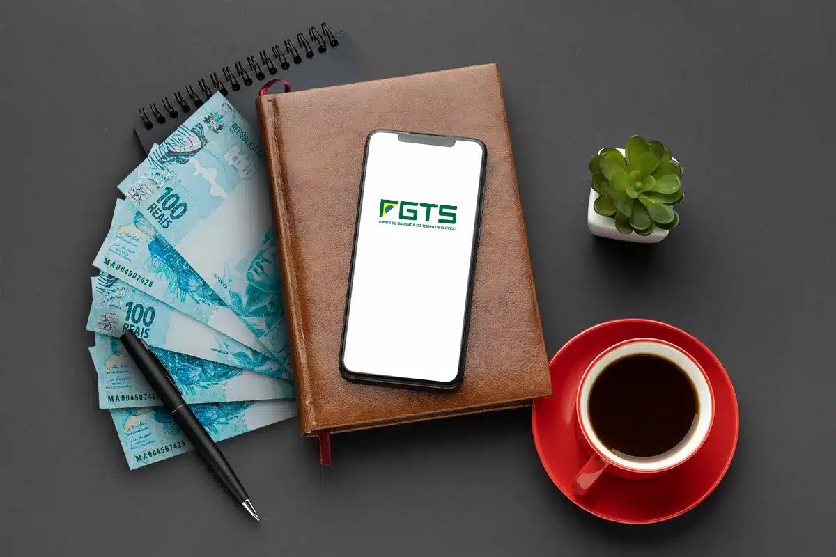 Celular exibindo logo do FGTS sobre caderno marrom, notas de R$ 100, caneta preta, xícara vermelha e suculenta.