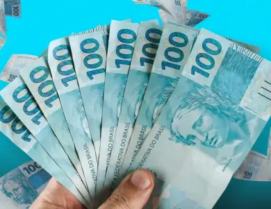 Mão segurando leque de notas de R$ 100 com fundo azul claro e cédulas espalhadas.