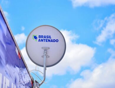Antena parabólica digital branca com logotipo "Brasil Antenado" instalada em estrutura azul contra céu azul com nuvens.